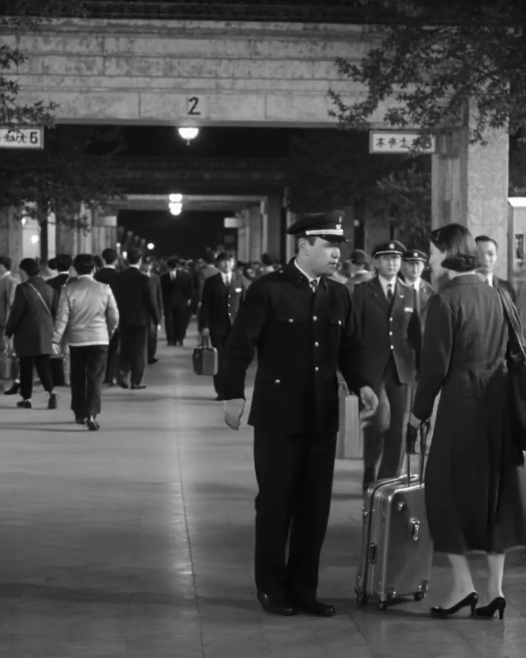 夜の駅のコンコースで、制服の駅員がスーツケースを持った外国人の女性旅行者と話している