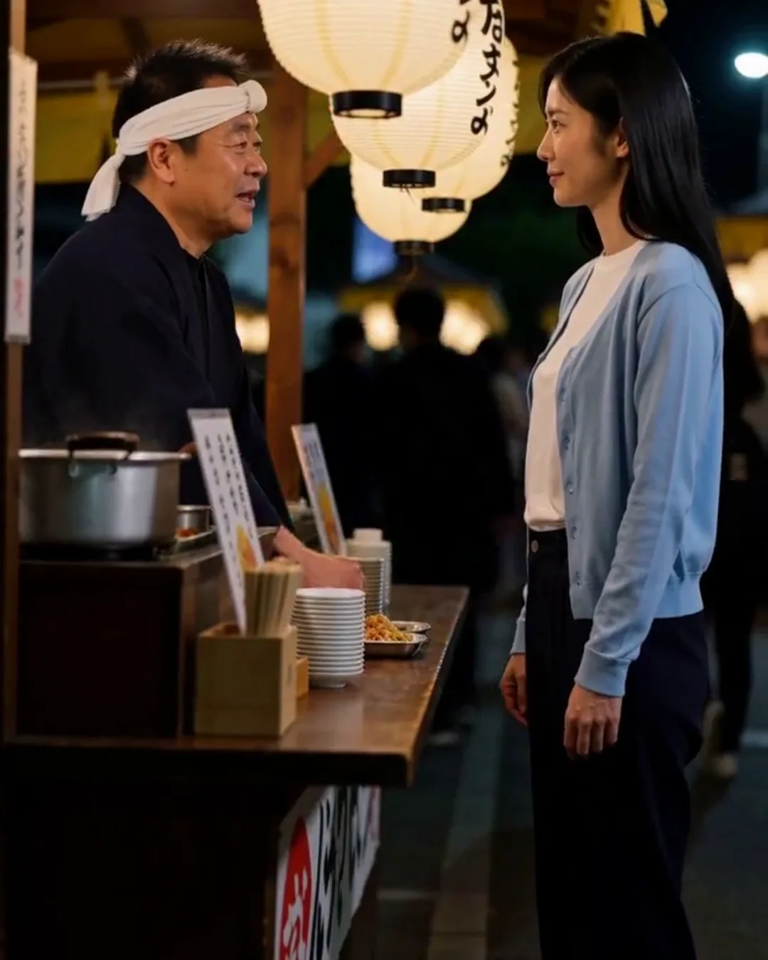 夜の屋台で、鉢巻きの店主と外国人の女性客が提灯の下で話している