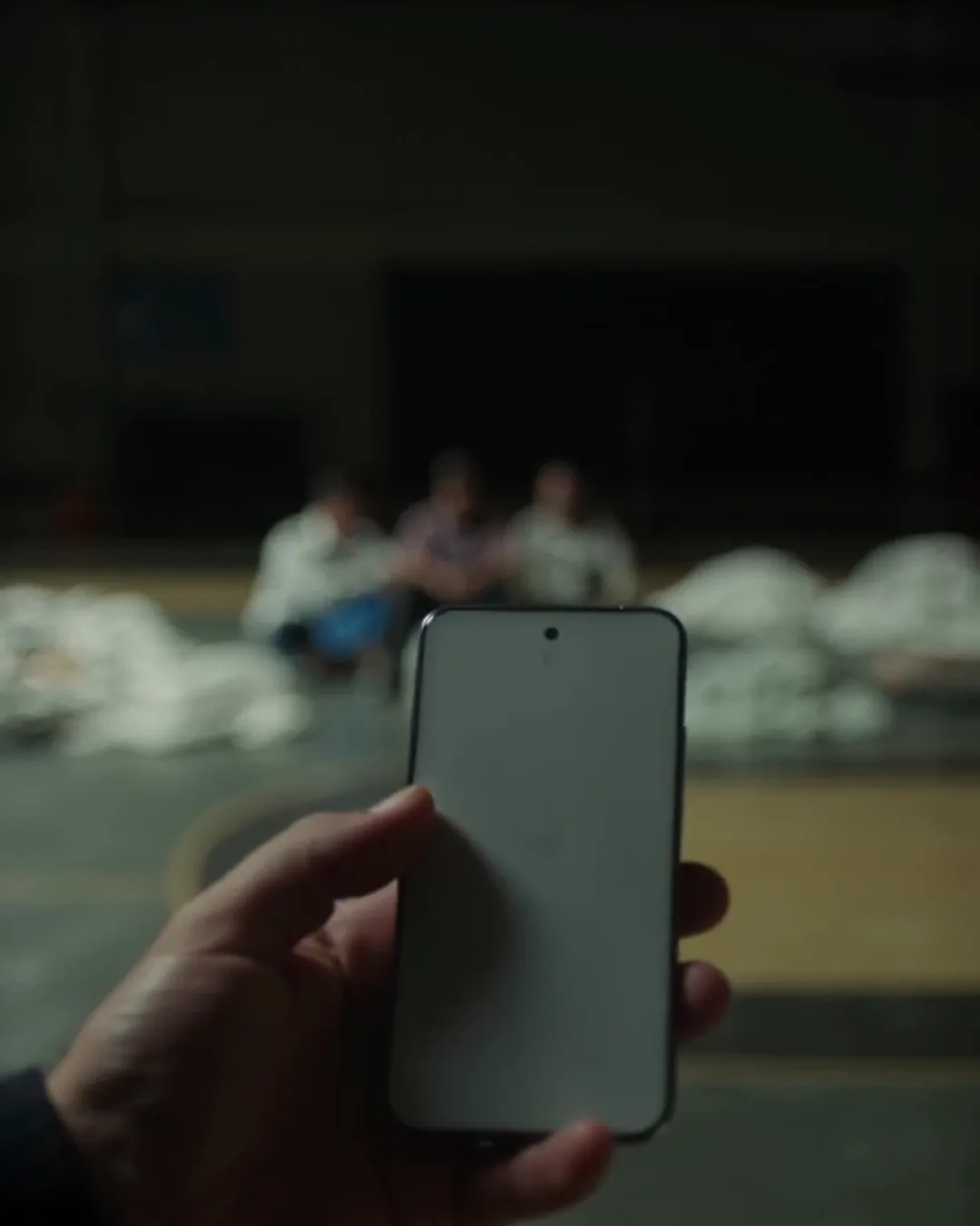 暗い体育館の中で、スマートフォンを持つ手。奥に毛布と座っている人がぼんやり見える