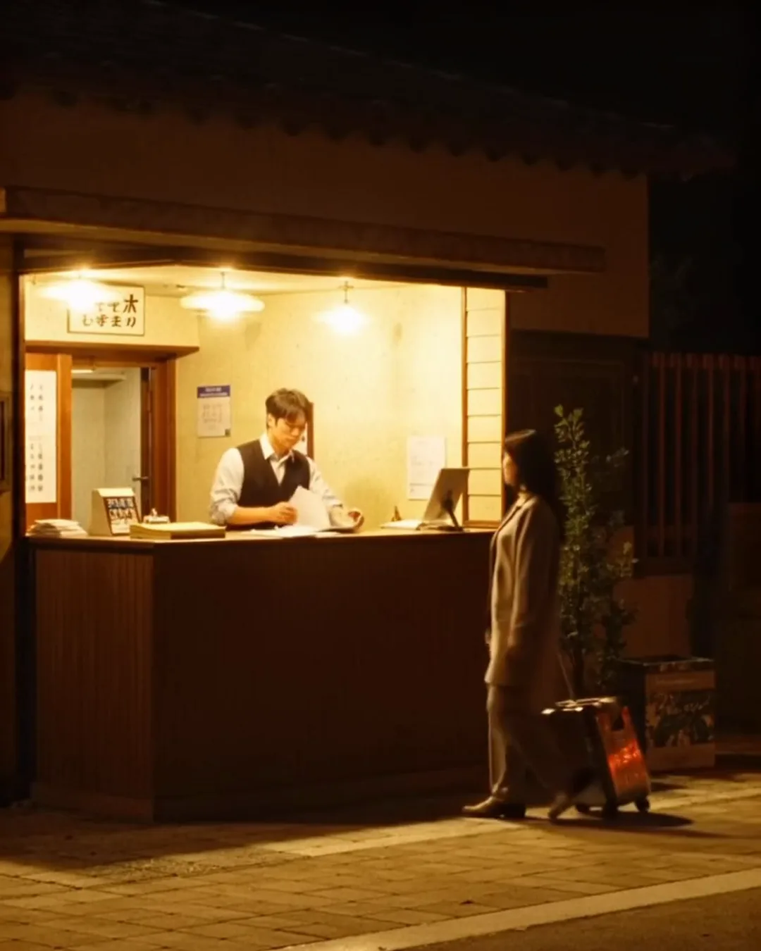 深夜のホテルのフロントで、ベスト姿の男性スタッフがスーツケースを持った外国人の女性客と話している