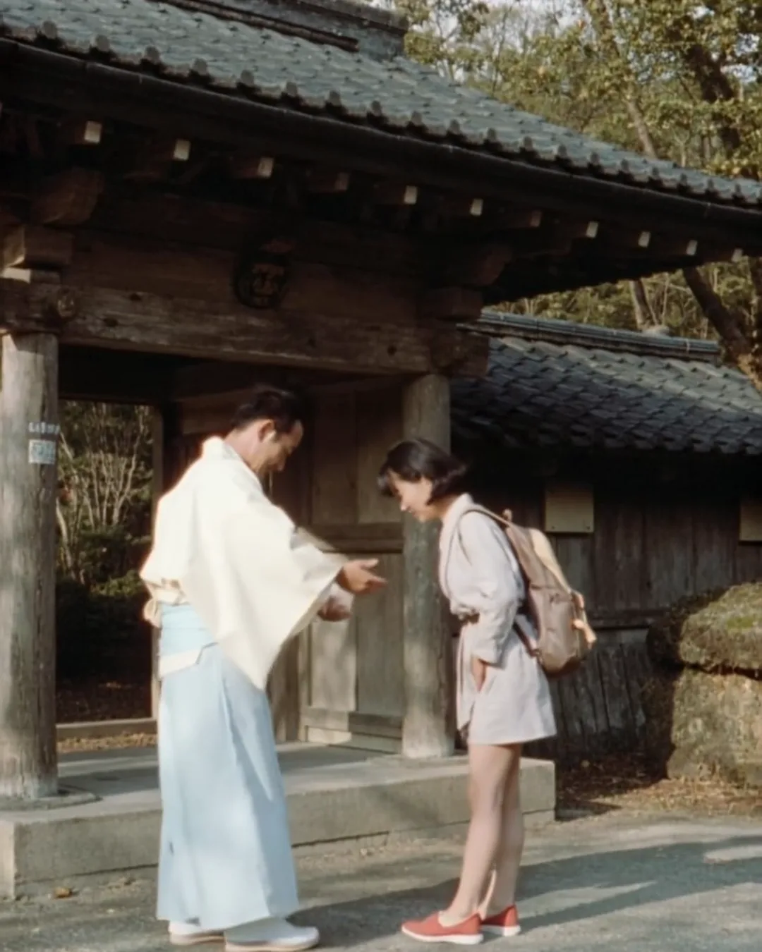 朝の神社の拝殿の前で、白い装束の神職が外国人の女性旅行者におじぎの仕方を示している